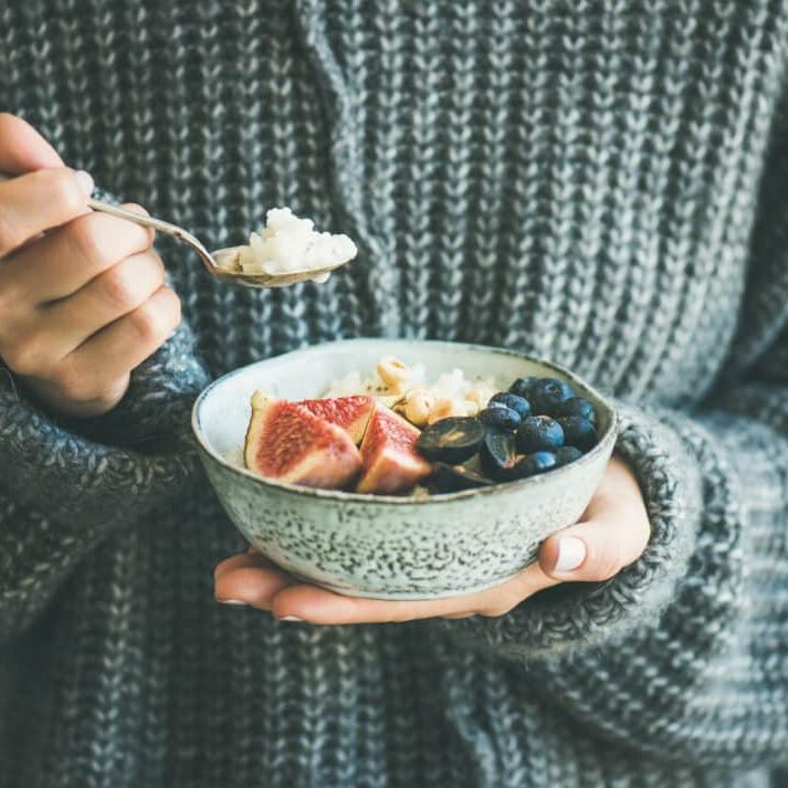 Healthy winter breakfast. Woman in woolen sweater eating rice coconut porridge with figs, berries, hazelnuts. Clean eating, vegetarian, vegan, alkiline diet food concept